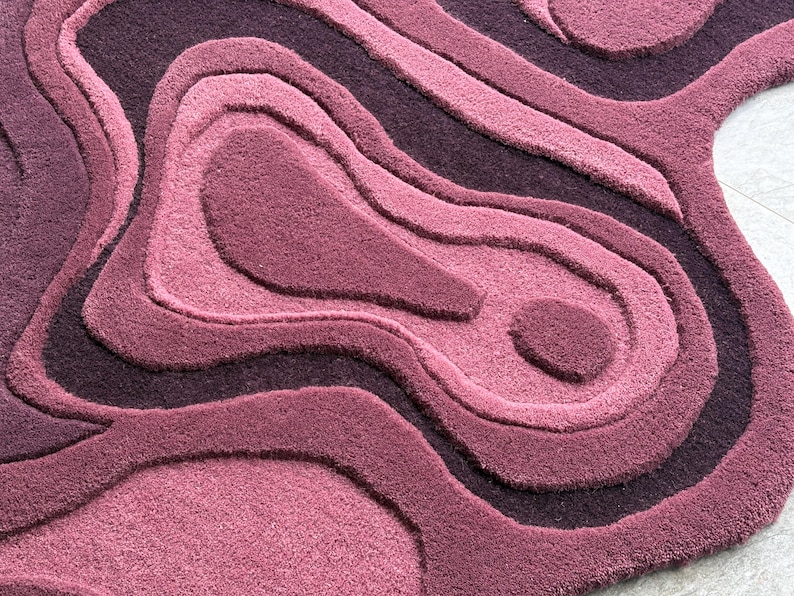 May include: A plush, abstract rug in shades of purple and mauve. The rug features a layered, topographic design with varying pile heights, creating a textured, three-dimensional effect. The rug's design resembles contour lines, adding visual interest to any space.