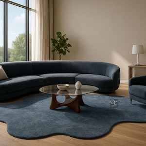 Pode incluir: Uma sala de estar moderna com um sof&aacute; curvo azul marinho, uma poltrona a condizer e um tapete com uma forma org&acirc;nica &uacute;nica. Uma mesa de centro com tampo de vidro e base de madeira est&aacute; no centro. A sala &eacute; preenchida com luz natural de uma grande janela.