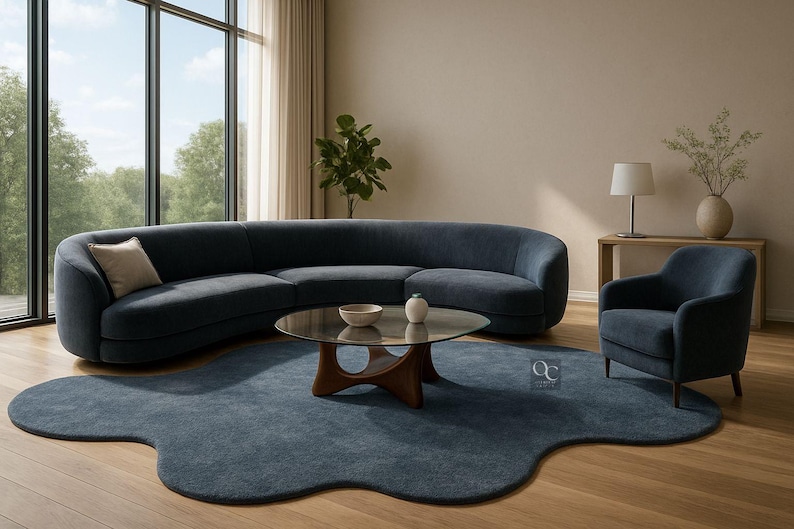 Pode incluir: Uma sala de estar moderna com um sof&aacute; curvo azul marinho, uma poltrona a condizer e um tapete com uma forma org&acirc;nica &uacute;nica. Uma mesa de centro com tampo de vidro e base de madeira est&aacute; no centro. A sala &eacute; preenchida com luz natural de uma grande janela.