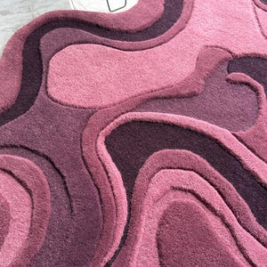 May include: A plush, abstract rug in shades of pink and purple. The rug features a layered, organic design with varying textures and tones, creating a visually dynamic and inviting surface. The color palette ranges from light rose to deep plum.
