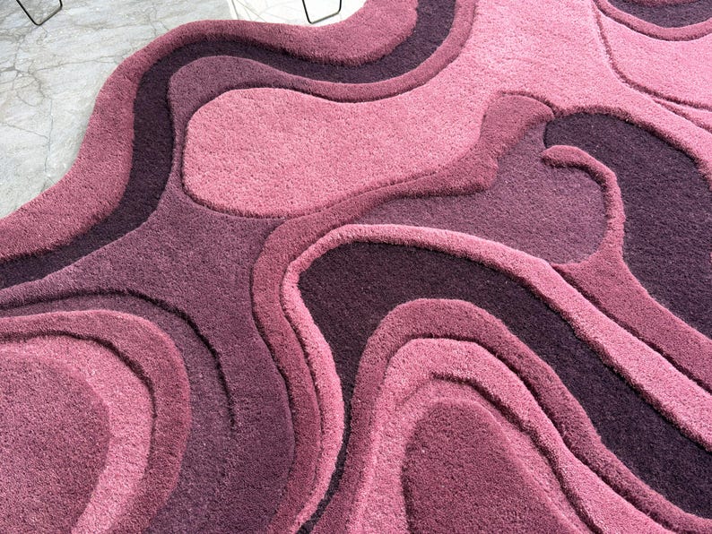 May include: A plush, abstract rug in shades of pink and purple. The rug features a layered, organic design with varying textures and tones, creating a visually dynamic and inviting surface. The color palette ranges from light rose to deep plum.