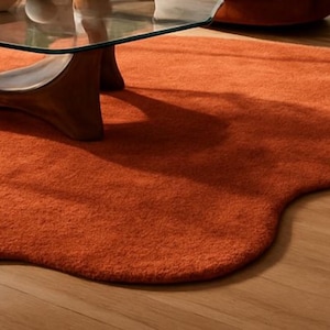 Pode incluir: Um tapete laranja queimado, de formato irregular e com superf&iacute;cie macia e texturizada. O tapete est&aacute; sobre um ch&atilde;o de madeira, com uma mesa de centro com tampo de vidro e uma cadeira laranja ao fundo. O design do tapete adiciona um toque moderno ao ambiente.