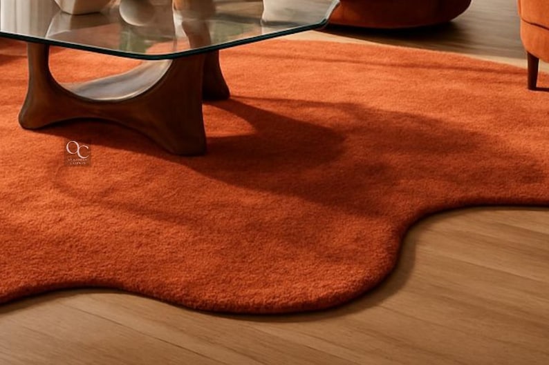 Pode incluir: Um tapete laranja queimado, de formato irregular e com superf&iacute;cie macia e texturizada. O tapete est&aacute; sobre um ch&atilde;o de madeira, com uma mesa de centro com tampo de vidro e uma cadeira laranja ao fundo. O design do tapete adiciona um toque moderno ao ambiente.