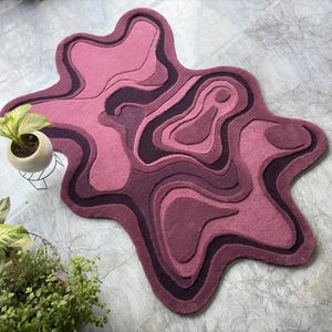 May include: A pink and purple abstract rug with a layered design. The rug has a unique, organic shape and is made of a soft, plush material. The rug is approximately 60 inches long and 40 inches wide.