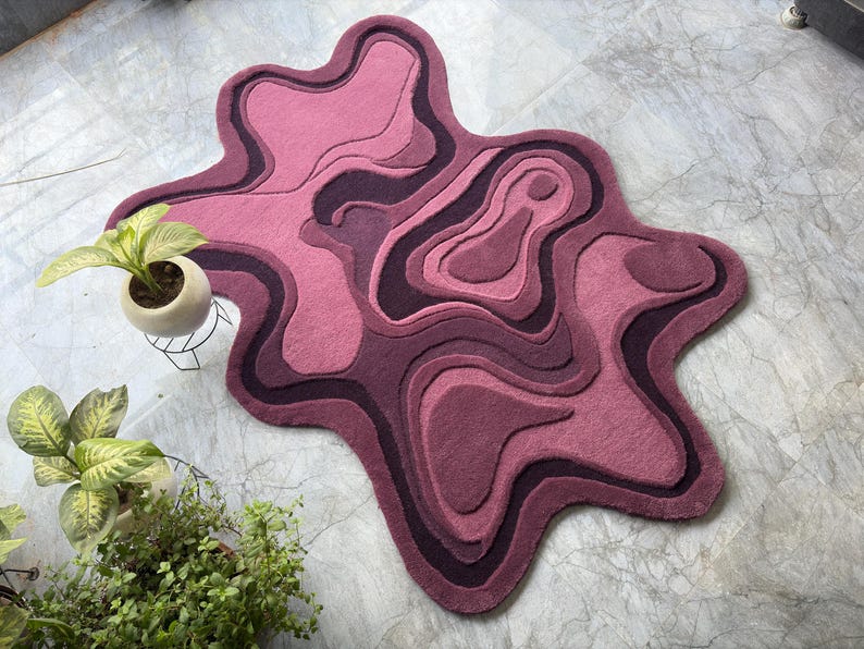 May include: A pink and purple abstract rug with a layered design. The rug has a unique, organic shape and is made of a soft, plush material. The rug is approximately 60 inches long and 40 inches wide.