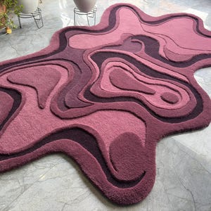 May include: A plush, abstract rug in shades of pink and purple. The rug features a layered, topographical design with varying depths and textures. The rug's unique shape and color palette create a modern, artistic statement for any room.