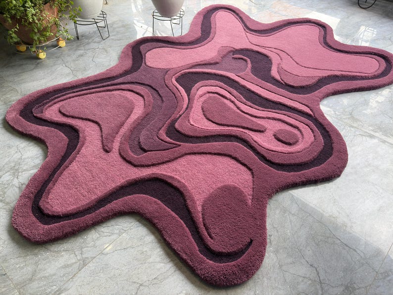 May include: A plush, abstract rug in shades of pink and purple. The rug features a layered, topographical design with varying depths and textures. The rug's unique shape and color palette create a modern, artistic statement for any room.