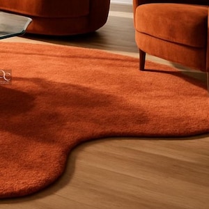 Pode incluir: Um tapete laranja, com formato org&acirc;nico e superf&iacute;cie macia e texturizada. O tapete est&aacute; colocado sobre um piso de madeira clara, com uma mesa com tampo de vidro e uma poltrona laranja vis&iacute;veis ao fundo. O formato &uacute;nico do tapete adiciona um toque moderno ao ambiente.
