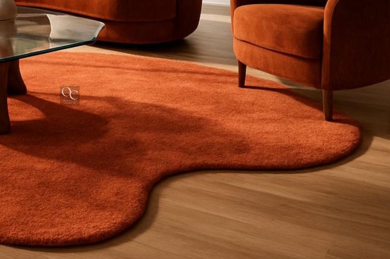 Pode incluir: Um tapete laranja, com formato org&acirc;nico e superf&iacute;cie macia e texturizada. O tapete est&aacute; colocado sobre um piso de madeira clara, com uma mesa com tampo de vidro e uma poltrona laranja vis&iacute;veis ao fundo. O formato &uacute;nico do tapete adiciona um toque moderno ao ambiente.