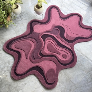 May include: A unique, abstract rug in shades of pink and purple. The rug features a layered design with a flowing, organic shape. The color palette includes various tones of pink and purple, creating a visually appealing and modern aesthetic.