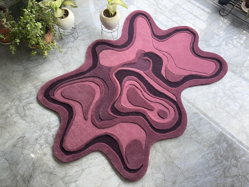 May include: A unique, abstract rug in shades of pink and purple. The rug features a layered design with a flowing, organic shape. The color palette includes various tones of pink and purple, creating a visually appealing and modern aesthetic.