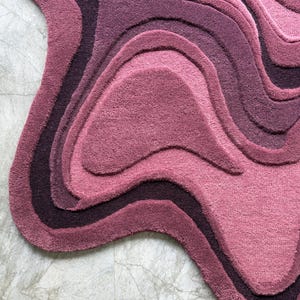 May include: A plush, abstract rug in shades of pink and purple. The rug features a layered design with wavy, organic shapes, creating a topographic effect. The color palette ranges from deep plum to light rose, offering a soft, textured appearance.