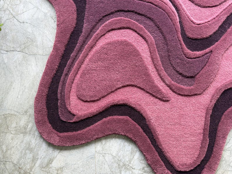 May include: A plush, abstract rug in shades of pink and purple. The rug features a layered design with wavy, organic shapes, creating a topographic effect. The color palette ranges from deep plum to light rose, offering a soft, textured appearance.
