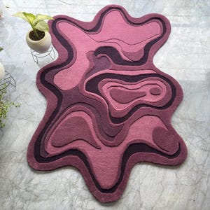May include: A unique, abstract-shaped rug in shades of pink and purple. The rug features a layered design with varying tones, creating a topographic effect. The rug's edges have a flowing, organic shape, adding to its modern aesthetic.
