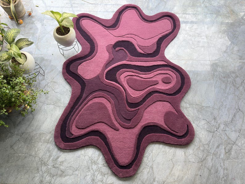 May include: A unique, abstract-shaped rug in shades of pink and purple. The rug features a layered design with varying tones, creating a topographic effect. The rug's edges have a flowing, organic shape, adding to its modern aesthetic.
