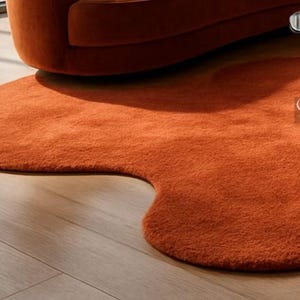 Pode incluir: Um tapete laranja, com formato org&acirc;nico e apar&ecirc;ncia macia e texturizada. Est&aacute; sobre um piso de madeira clara, com parte de um sof&aacute; laranja ao fundo. O design do tapete adiciona um toque moderno ao ambiente.