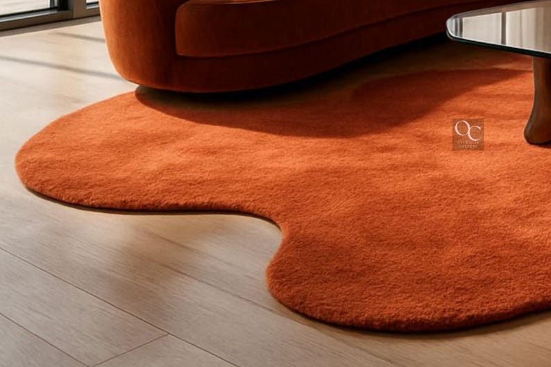 Pode incluir: Um tapete laranja, com formato org&acirc;nico e apar&ecirc;ncia macia e texturizada. Est&aacute; sobre um piso de madeira clara, com parte de um sof&aacute; laranja ao fundo. O design do tapete adiciona um toque moderno ao ambiente.