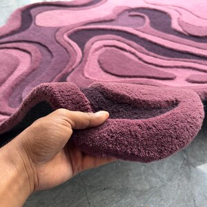 May include: A plush, abstract rug in shades of purple and pink. The rug features a layered, topographic design with varying textures and depths. The rug is being held up, revealing its thick, textured backing.