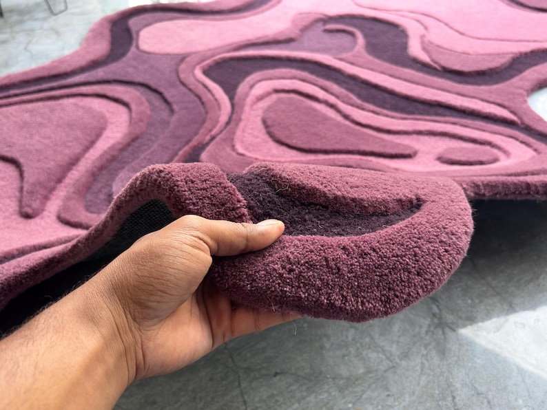 May include: A plush, abstract rug in shades of purple and pink. The rug features a layered, topographic design with varying textures and depths. The rug is being held up, revealing its thick, textured backing.