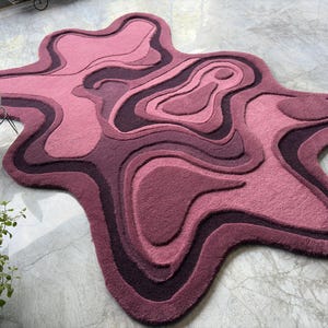 May include: A unique, abstract rug in shades of pink and purple. The rug features a layered, organic design with varying textures and depths. The rug's shape is irregular, resembling a topographical map. The rug is a decorative floor covering.