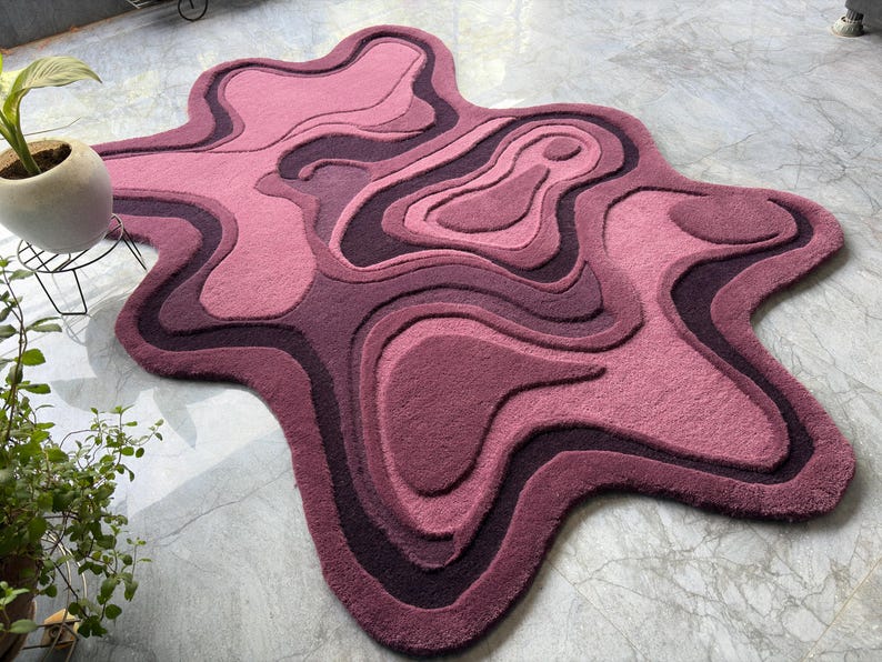 May include: A unique, abstract rug in shades of pink and purple. The rug features a layered, organic design with varying textures and depths. The rug's shape is irregular, resembling a topographical map. The rug is a decorative floor covering.