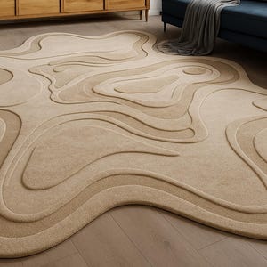 May include: Beige area rug with a unique, organic shape. The rug features a layered design with varying shades of beige, creating a topographic effect. The rug is placed on a wooden floor, with furniture visible in the background.