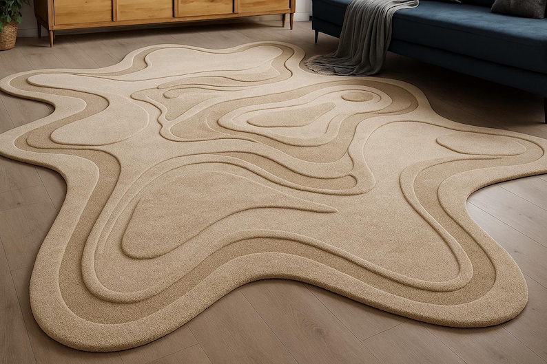 May include: Beige area rug with a unique, organic shape. The rug features a layered design with varying shades of beige, creating a topographic effect. The rug is placed on a wooden floor, with furniture visible in the background.