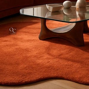 Pode incluir: Um tapete laranja com uma forma org&acirc;nica &uacute;nica. O tapete &eacute; feito de um material macio e texturizado e &eacute; colocado sob uma mesa de centro com tampo de vidro e base de madeira escura. O tapete adiciona calor e cor &agrave; sala.