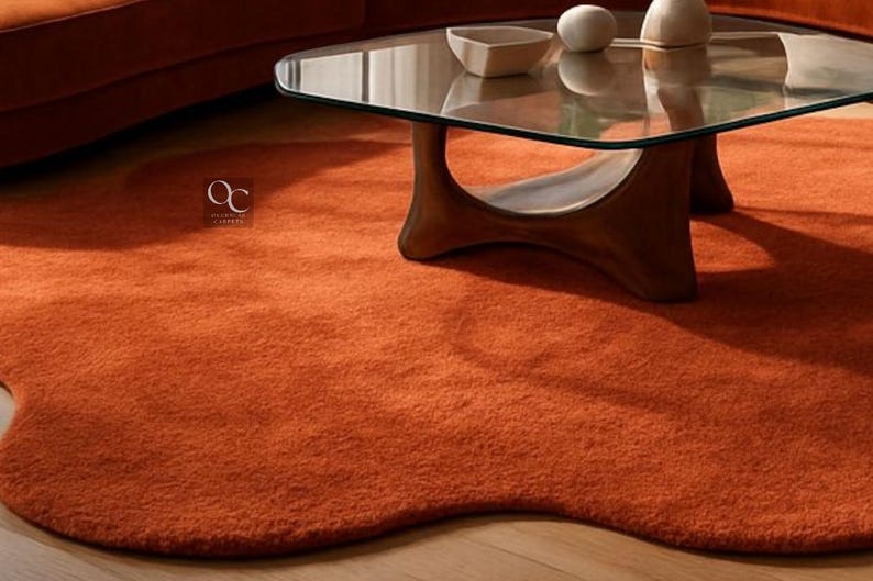 Pode incluir: Um tapete laranja com uma forma org&acirc;nica &uacute;nica. O tapete &eacute; feito de um material macio e texturizado e &eacute; colocado sob uma mesa de centro com tampo de vidro e base de madeira escura. O tapete adiciona calor e cor &agrave; sala.