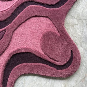 May include: A plush, abstract rug in shades of mauve, plum, and deep purple. The rug features a layered, wave-like design with varying textures and depths. The rug's edge has a unique, organic shape.