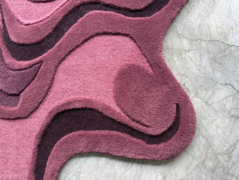 May include: A plush, abstract rug in shades of mauve, plum, and deep purple. The rug features a layered, wave-like design with varying textures and depths. The rug's edge has a unique, organic shape.