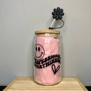 Könnte beinhalten: Ein klares Glasgefäß mit einem Bambusdeckel und einem schwarzen strohhalm in Form einer Blume. Das Gefäß enthält ein rosa Taschentuch und eine schwarze Grafik mit der Aufschrift "Professional Overthinker" mit einem Smiley und einem Herz.