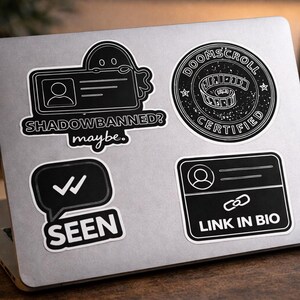 Puede incluir: Un conjunto de pegatinas en blanco y negro sobre un portátil. Las pegatinas incluyen una tarjeta de identificación con forma de fantasma con el texto "SHADOWBANNED? maybe.", un diseño circular "DOOMSCROLL CERTIFIED", una burbuja de diálogo con marcas de verificación y la palabra "SEEN", y una tarjeta de identificación con un icono de enlace y el texto "LINK IN BIO".