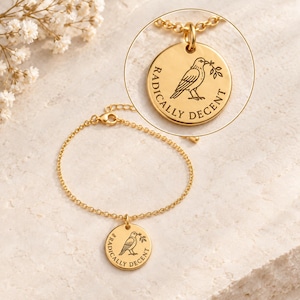 May include: A gold-toned chain bracelet featuring a circular pendant engraved with a bird holding a sprig and the text RADICALLY DECENT. The design is shown in full and in a magnified circular inset, highlighting the detailed engraving on the metal.