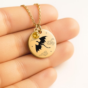 May include: A gold-toned circular pendant necklace featuring a black silhouette of a flying dragon, engraved clouds, and small stars. A small, round, yellow faceted gemstone is attached to the top of the disc, suspended from a gold-toned chain.