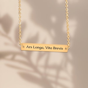 Peut inclure: Un collier doré avec un pendentif rectangulaire. Le pendentif porte l'inscription "Ars Longa, Vita Brevis" en noir. La chaîne du collier est fine et également dorée.