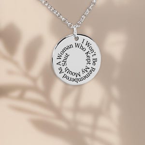 Puede incluir: Un collar con colgante plateado con un dije circular. El dije tiene el texto grabado: "Como mujer, no ser&eacute; recordada quien mantuvo la boca cerrada."