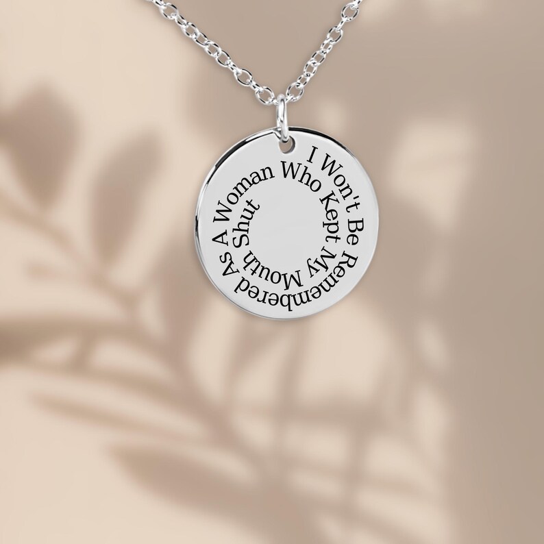 Puede incluir: Un collar con colgante plateado con un dije circular. El dije tiene el texto grabado: "Como mujer, no ser&eacute; recordada quien mantuvo la boca cerrada."