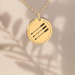 Könnte beinhalten: Goldfarbene Halskette mit Anhänger, die einen kreisförmigen Anhänger mit drei schwarzen Pfeilen zeigt, die diagonal nach unten zeigen. Die Pfeile sind parallel angeordnet. Die Halskette ist ebenfalls goldfarben.