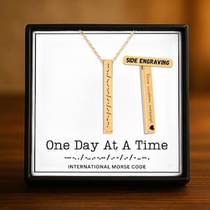 May include: A gold-colored vertical bar necklace with Morse code, presented in a black box. The necklace has the phrase "One Day At A Time" in Morse code. The box also includes a side engraving option with the text "Your custom message".