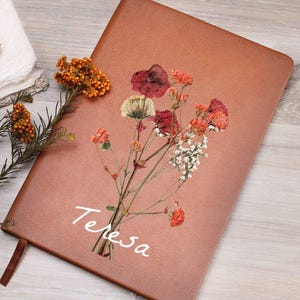 May include: A brown leather journal with the name "Teresa" in white script. The journal is decorated with a bouquet of dried flowers in shades of red, orange, and white. A brown ribbon bookmark is visible.