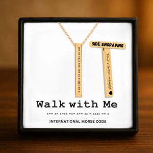 Könnte beinhalten: Goldfarbene Halskette mit vertikalem Anhänger mit Morsecode. Die Halskette wird in einer schwarzen Box mit den Worten "Walk with Me" und "International Morse Code" präsentiert. Ein separater Balken zeigt "Side Engraving".