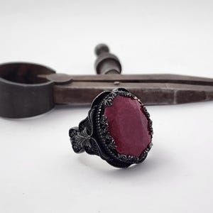 Może przedstawiać: Srebrny pierścionek z dużym, owalnym, ciemnoczerwonym kamieniem szlachetnym osadzonym w misternym, czarnym wzorze filigranowym.