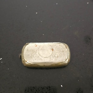 May include: A small, rectangular, silver-toned metal case with rounded edges. The case has a hinged lid and a circular design on the top. The metal shows signs of wear and age, with some tarnishing and discoloration.