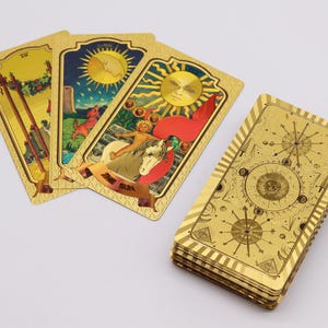 May include: A deck of gold tarot cards with detailed illustrations. The cards feature images of the sun, moon, and other celestial bodies. The card backs have a geometric design. The cards are arranged in a fan shape.