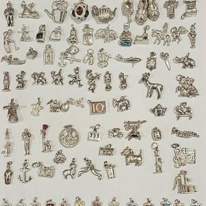 May include: A large collection of assorted silver-toned metal charms featuring various shapes including animals, vehicles, buildings, musical instruments, household items, and a row of small colourful heraldic crest shields at the bottom.