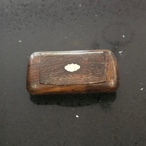May include: A small, rectangular wooden box with a hinged lid. The box is a dark brown colour with a decorative, light-coloured inlay on the lid. The box shows signs of age, with wear and tear visible on the wood, suggesting it is antique.