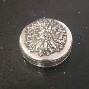 May include: A small, circular, silver-coloured box featuring a detailed floral design on the lid. The flower is in high relief, with intricate petal detailing. The box has a simple, elegant design, suitable for jewellery or small keepsakes.