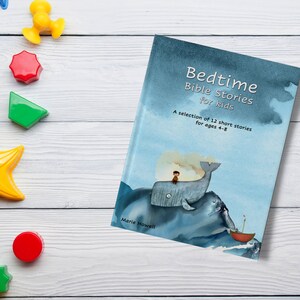 Op de afbeelding: Een blauw boek met de titel "Bedtime Bible Stories for Kids" en een aquarel-illustratie van een walvis met een persoon erop. Het boek ligt op een wit houten oppervlak met kleurrijke speelgoed.