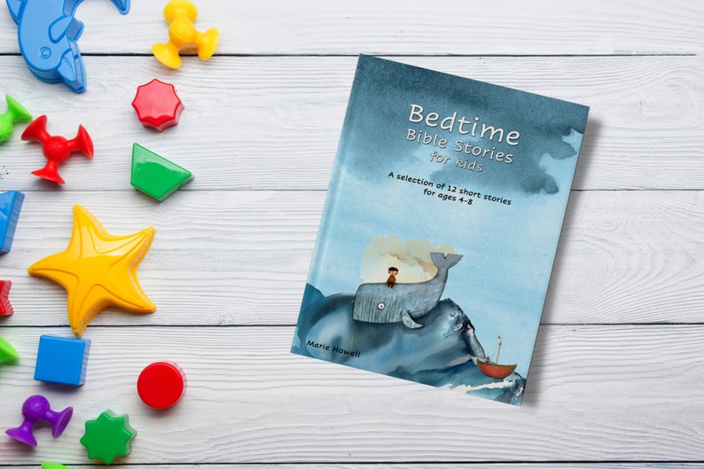 Op de afbeelding: Een blauw boek met de titel "Bedtime Bible Stories for Kids" en een aquarel-illustratie van een walvis met een persoon erop. Het boek ligt op een wit houten oppervlak met kleurrijke speelgoed.